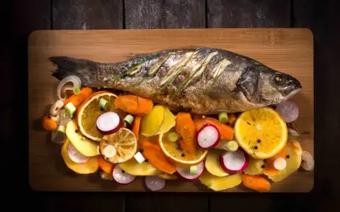 Eine ganze Forelle im Backofen, serviert mit bunten Gemüsescheiben wie Karotten, Radieschen und Orangen auf einem Holzbrett.