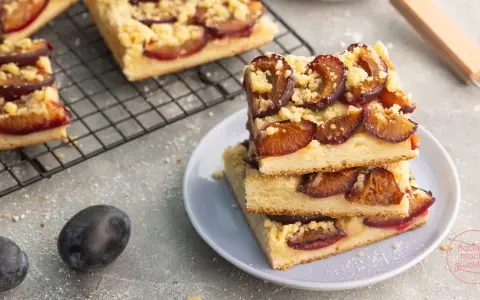 Saftiger zwetschgenkuchen vegan mit Streuseln auf einem Teller gestapelt. Daneben frische Zwetschgen und ein Kuchenblech.