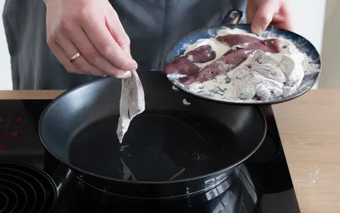 Ein Stück Leber wird in eine heiße Pfanne mit Öl gelegt, um sie zu braten. Auf einem Teller liegen weitere Leberstücke, die in Mehl gewendet wurden.