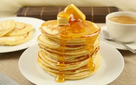 Ein Stapel goldbrauner Pancakes, übergossen mit Honig und Butter, daneben eine Tasse Kaffee. Perfekt für ein leckeres Pancakes Rezept.