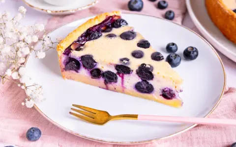 Ein Stück Heidelbeerkuchen Landfrauenart mit saftigen Blaubeeren und cremigem Guss auf einem Teller, daneben eine goldene Gabel und frische Beeren.
