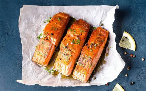 Drei saftige **Lachs im Ofen**-Portionen, garniert mit Kräutern und Pfeffer, auf Backpapier. Daneben Zitronenspalten.