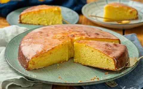 Ein saftiger Zitronenkuchen mit glänzender Glasur, auf einem grünen Teller serviert. Ein Stück wird gerade herausgehoben.