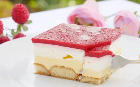 Himbeer-Sahne-Schnitten auf weißer Platte, mit frischen Himbeeren und rosa Rosen im Hintergrund. Ein Stück wird mit einer Gabel herausgenommen.