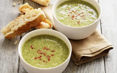 Zwei Schalen cremiger Brokkolisuppe, garniert mit roten Chiliflocken, stehen neben knusprigem Baguette auf einem Holztisch.