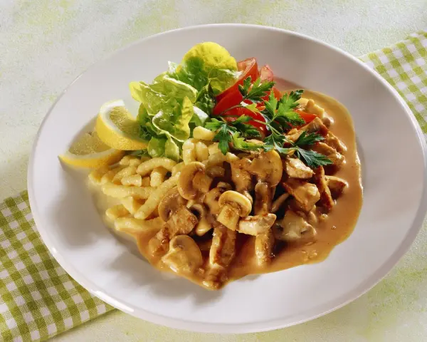 Ein Teller mit geschnetzeltem Fleisch mit Pilzen in cremiger Sauce, dazu Spätzle, Salat und Zitronenspalten.