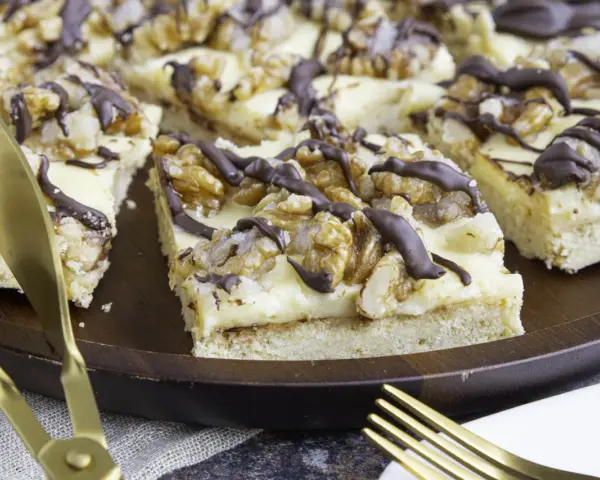 Ein "friss dich dumm kuchen" mit cremiger Füllung, Walnüssen und Schokoladendrizzle auf einem Holztablett. Goldene Gabel und Messer liegen daneben.
