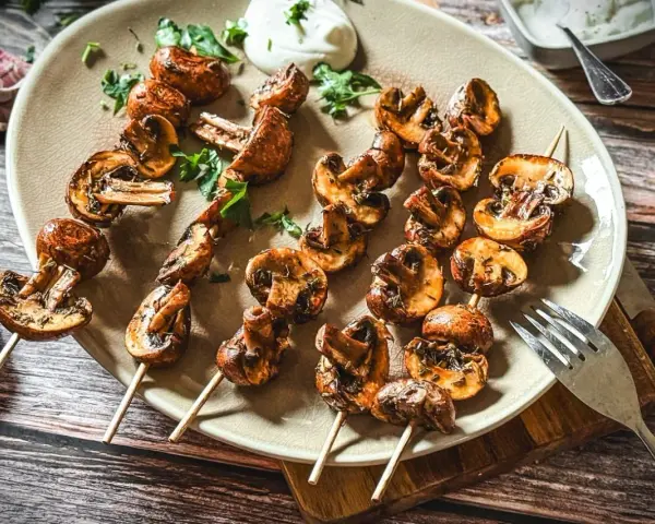 Gegrillte Champignons auf Spießen, mit Kräutern verfeinert und auf einem Teller mit Dip serviert. Ein Genuss für jeden Pilzliebhaber!