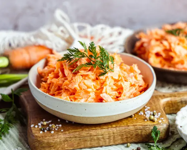 Karottensalat mit cremigem Dressing, wie im Restaurant. Frisch geriebene Karotten, garniert mit Dill, auf einem Holzbrett.