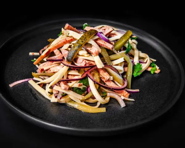 Ein appetitlicher Schweizer Wurstsalat mit Wurststreifen, Käse, roten Zwiebeln und Gewürzgurken auf einem dunklen Teller.