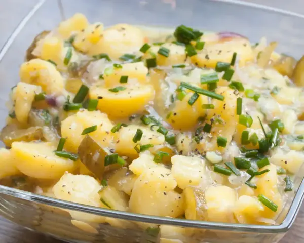 Ein köstlicher Kartoffelsalat ohne Mayo, mit Gurkenstücken und frischem Schnittlauch garniert, in einer Glasschüssel.
