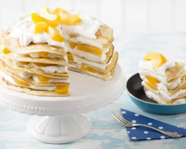 Eine köstliche Pfannkuchen Torte mit Pfirsichen und Sahne, auf einem weißen Tortenständer und ein Stück in einer kleinen Pfanne.