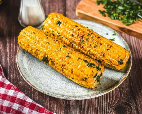 Zwei goldgelbe maiskolben, perfekt gegrillt und mit Kräutern bestreut, liegen auf einem Teller. Ein Genuss für alle, die maiskolben grillen lieben.