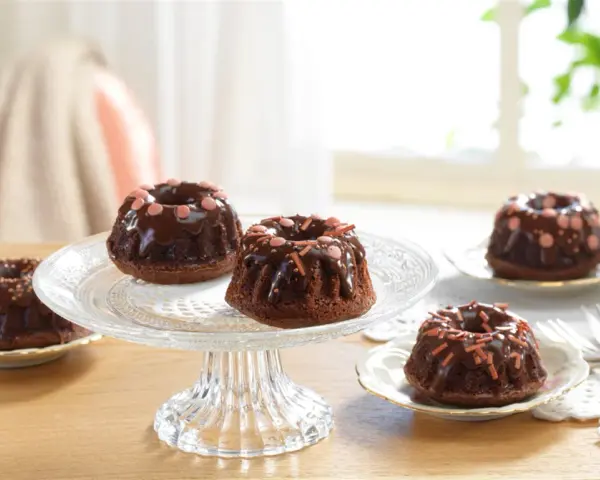Schokoladen-Mini-Gugelhupf dekorieren mit Schokoglasur und bunten Streuseln auf einem Tortenständer.