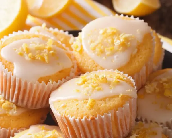 Leckere zitronenmuffins rezept: goldgelbe Muffins mit weißer Glasur und Zitronenschale bestreut, daneben frische Zitronenscheiben.