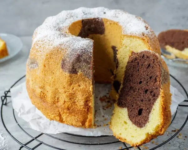 Ein saftiger Marmorkuchen Gugelhupf, mit Puderzucker bestäubt, wartet auf seinen Anschnitt. Ein Stück ist bereits abgetrennt.