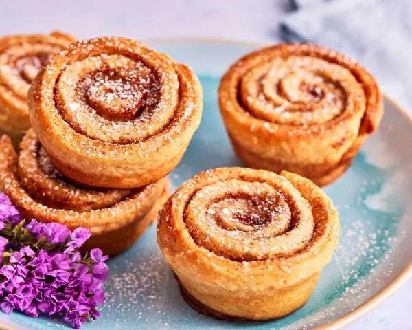 Saftige Zimtschnecken Muffins, bestäubt mit Puderzucker, liegen auf einem blauen Teller. Lila Blumen schmücken die süße Leckerei.