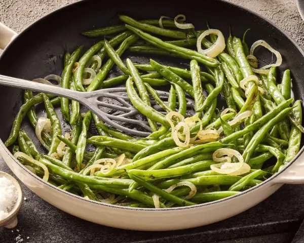 Grüne Bohnen mit Zwiebelringen in einer Pfanne, bereit zum Kochen. Daneben eine Schale mit grobem Salz.