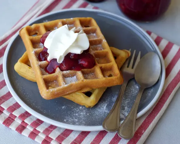 Zwei Hefewaffeln mit Trockenhefe, garniert mit Kirschkompott und Sahne, bestäubt mit Puderzucker.