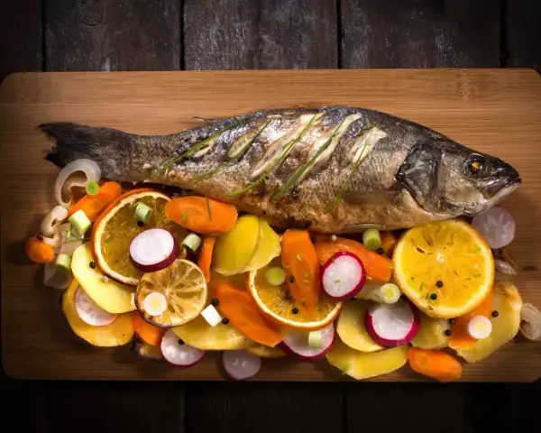 Eine ganze Forelle im Backofen, serviert mit bunten Gemüsescheiben wie Karotten, Radieschen und Orangen auf einem Holzbrett.