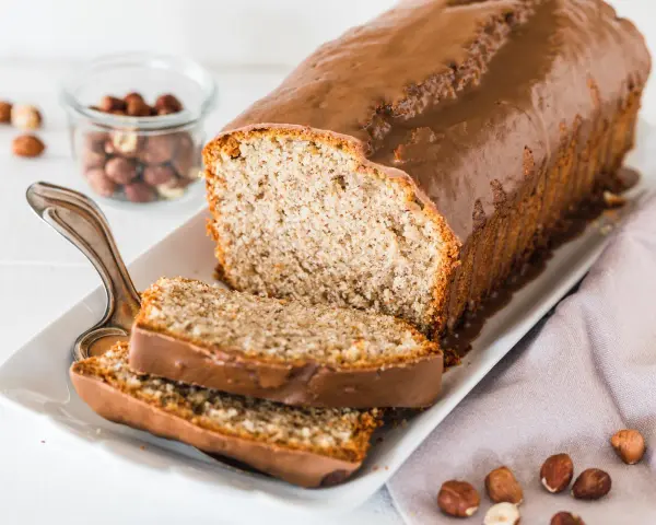Saftiger Mohn Nuss Kuchen mit Schokoladenglasur, daneben ein Glas mit Haselnüssen und zwei angeschnittene Scheiben.