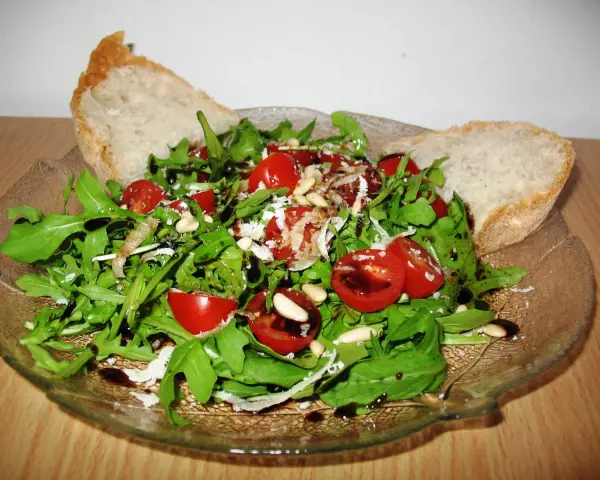 Frischer Rucola Salat mit Kirschtomaten, Pinienkernen und Parmesan, beträufelt mit Balsamico-Dressing. Dazu knuspriges Brot.