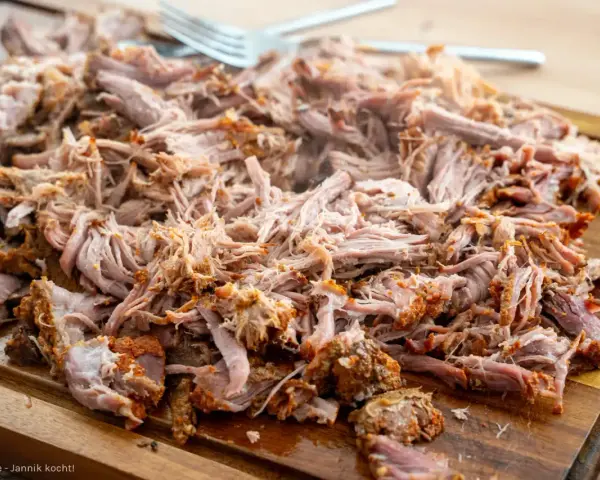 Ein Haufen zartes, **gezupftes Fleisch** liegt auf einem Holzbrett, bereit für leckere Sandwiches oder Tacos. Eine Gabel liegt daneben.