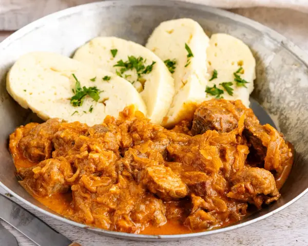 Herzhaftes Szegediner Gulasch mit Klößen, garniert mit Petersilie, serviert in einer Metallschüssel.