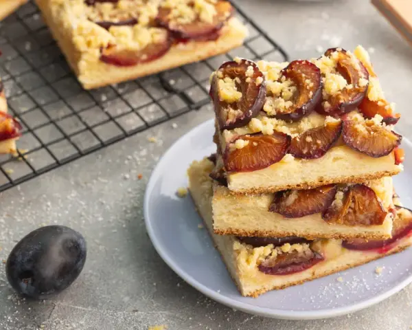Saftiger zwetschgenkuchen vegan mit Streuseln auf einem Teller gestapelt. Daneben frische Zwetschgen und ein Kuchenblech.