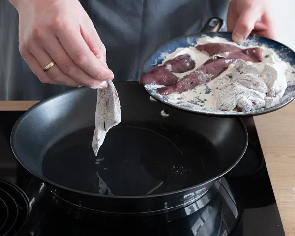 Ein Stück Leber wird in eine heiße Pfanne mit Öl gelegt, um sie zu braten. Auf einem Teller liegen weitere Leberstücke, die in Mehl gewendet wurden.