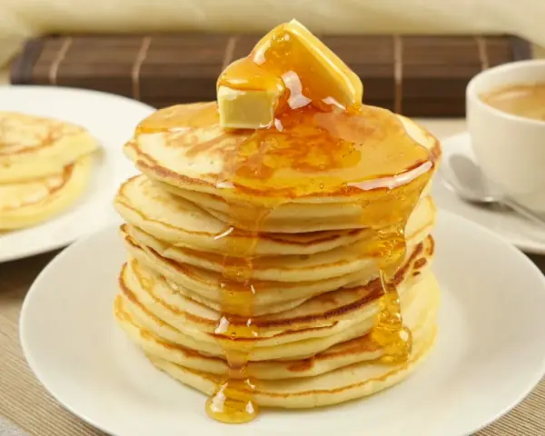 Ein Stapel goldbrauner Pancakes, übergossen mit Honig und Butter, daneben eine Tasse Kaffee. Perfekt für ein leckeres Pancakes Rezept.