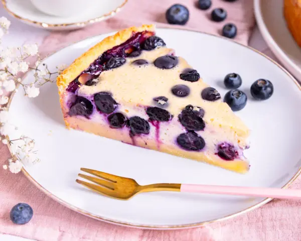 Ein Stück Heidelbeerkuchen Landfrauenart mit saftigen Blaubeeren und cremigem Guss auf einem Teller, daneben eine goldene Gabel und frische Beeren.
