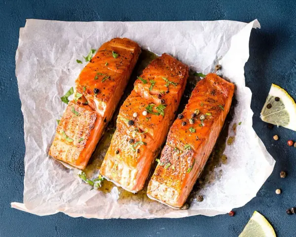Drei saftige **Lachs im Ofen**-Portionen, garniert mit Kräutern und Pfeffer, auf Backpapier. Daneben Zitronenspalten.