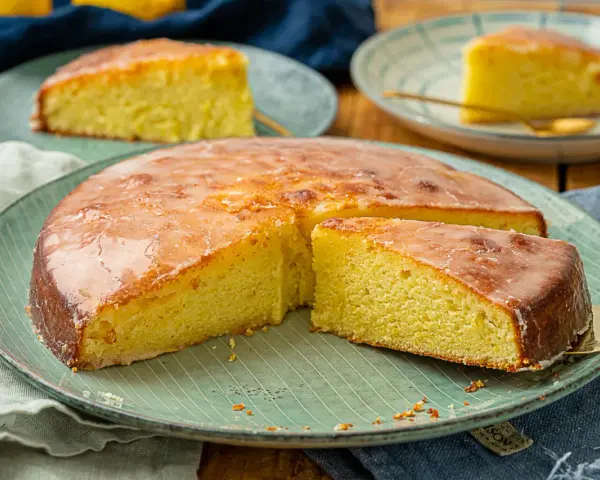 Ein saftiger Zitronenkuchen mit glänzender Glasur, auf einem grünen Teller serviert. Ein Stück wird gerade herausgehoben.