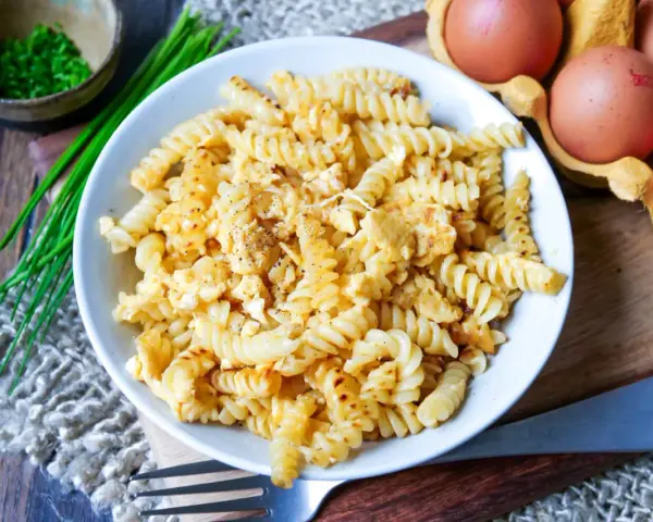 Fusilli-Nudeln mit Rührei, bestreut mit Pfeffer. Daneben liegen frische Eier und Schnittlauch. Ein einfaches Gericht: Nudeln mit Ei.