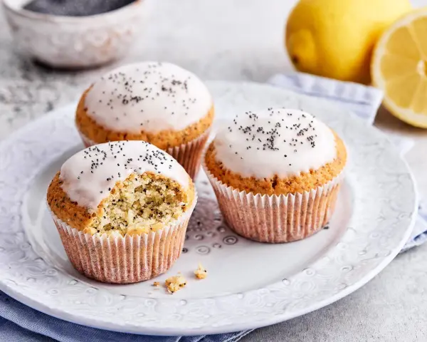Drei köstliche zitronen mohn muffins mit Zuckerguss und Mohn bestreut, daneben frische Zitronen.