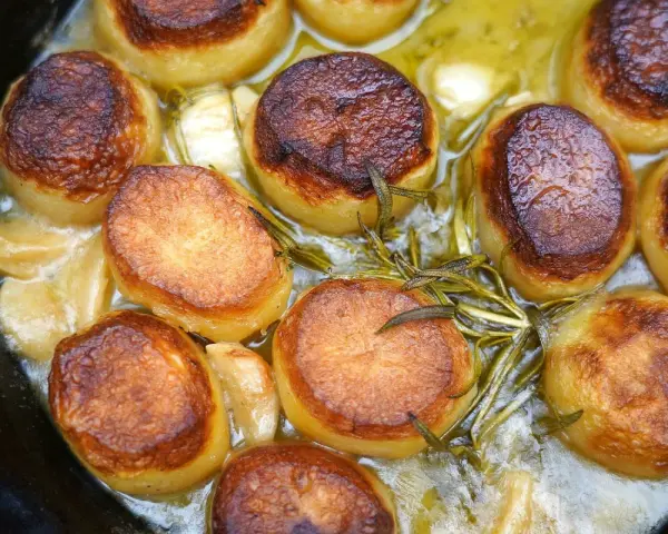 Goldbraun gebratene Fondant-Kartoffeln mit Rosmarin und Knoblauch in einer Pfanne.