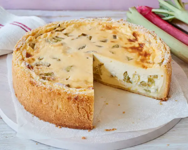 Ein saftiger Rhabarberkuchen, schnell gebacken und mit Puderzucker bestäubt. Daneben liegen frische Rhabarberstangen.