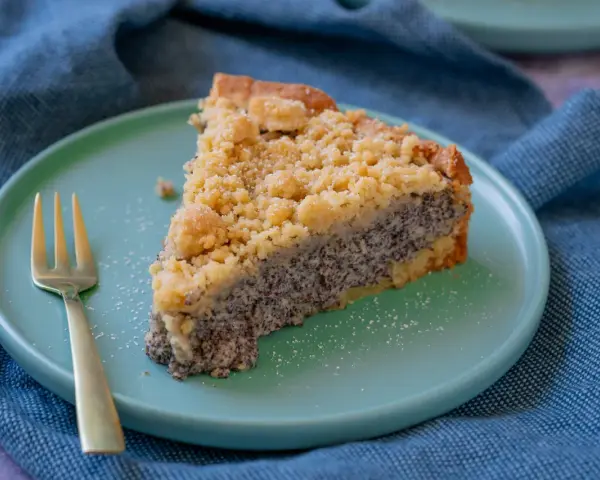 Ein saftiges Stück Mohnkuchen mit Streuseln auf einem mintgrünen Teller, daneben eine goldene Gabel. Ein köstliches Mohnkuchen Rezept.