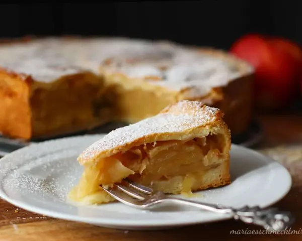 Ein Stück gedeckter Apfelkuchen mit Puderzucker bestäubt, auf einem Teller mit einer Gabel. Im Hintergrund der ganze Kuchen und Äpfel.