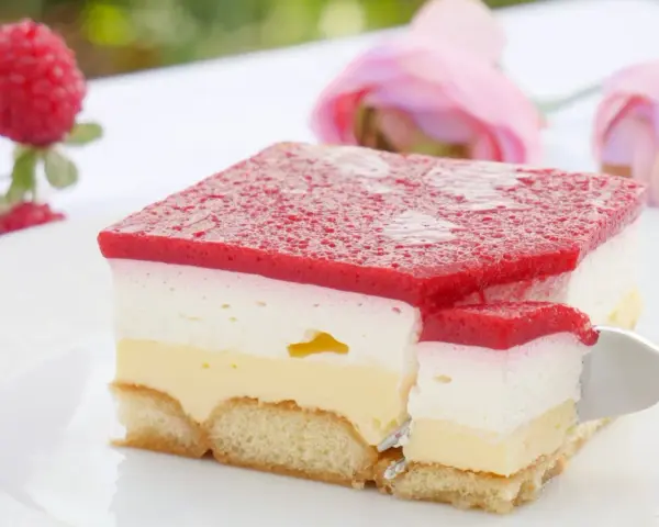 Himbeer-Sahne-Schnitten auf weißer Platte, mit frischen Himbeeren und rosa Rosen im Hintergrund. Ein Stück wird mit einer Gabel herausgenommen.