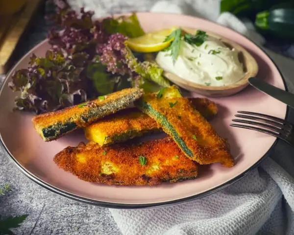 Knuspriges Zucchini-Schnitzel auf rosa Teller, serviert mit Dip, Salat und Zitrone. Ein Genuss für jeden Tag!