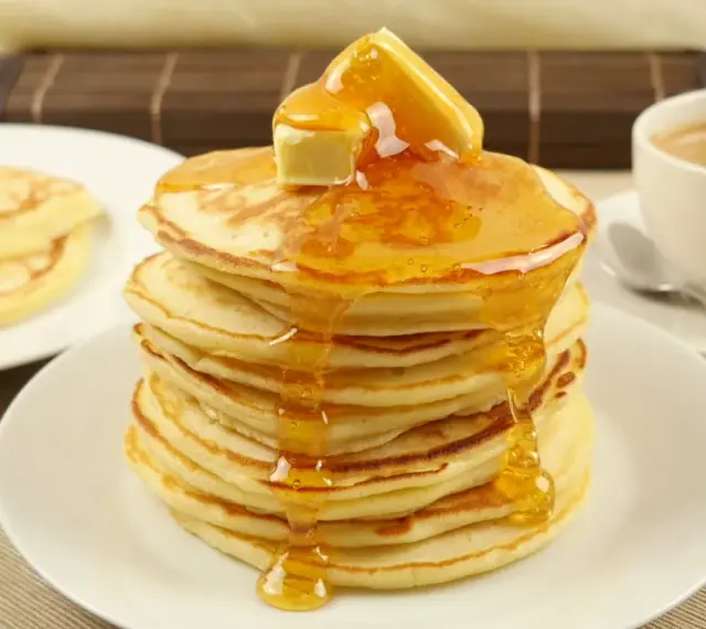Ein Stapel goldbrauner Pancakes, übergossen mit Honig und Butter, daneben eine Tasse Kaffee. Perfekt für ein leckeres Pancakes Rezept.