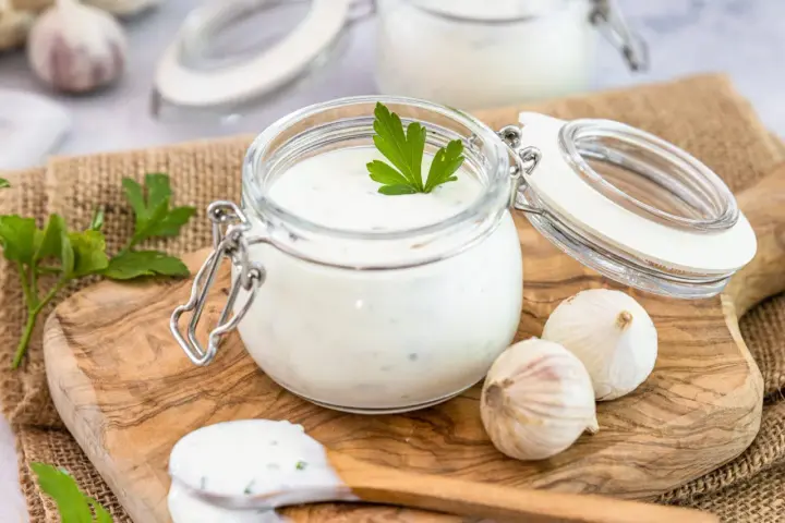 Ein Glas mit cremigem Knoblauch Dip, garniert mit Petersilie, daneben Knoblauchzehen und ein Löffel mit Dip.