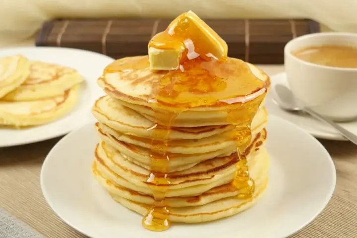Ein Stapel goldbrauner Pancakes, übergossen mit Honig und Butter, daneben eine Tasse Kaffee. Perfekt für ein leckeres Pancakes Rezept.