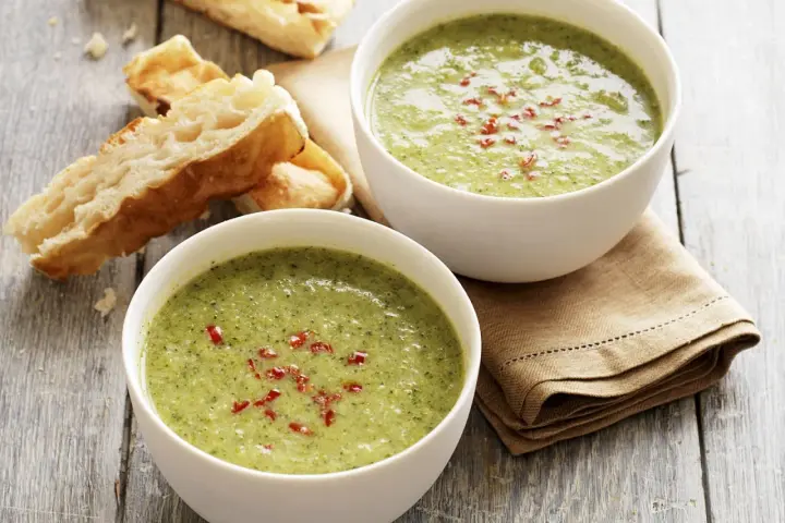 Zwei Schalen cremiger Brokkolisuppe, garniert mit roten Chiliflocken, stehen neben knusprigem Baguette auf einem Holztisch.