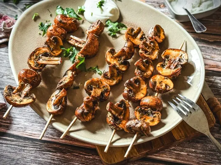 Gegrillte Champignons auf Spießen, mit Kräutern verfeinert und auf einem Teller mit Dip serviert. Ein Genuss für jeden Pilzliebhaber!