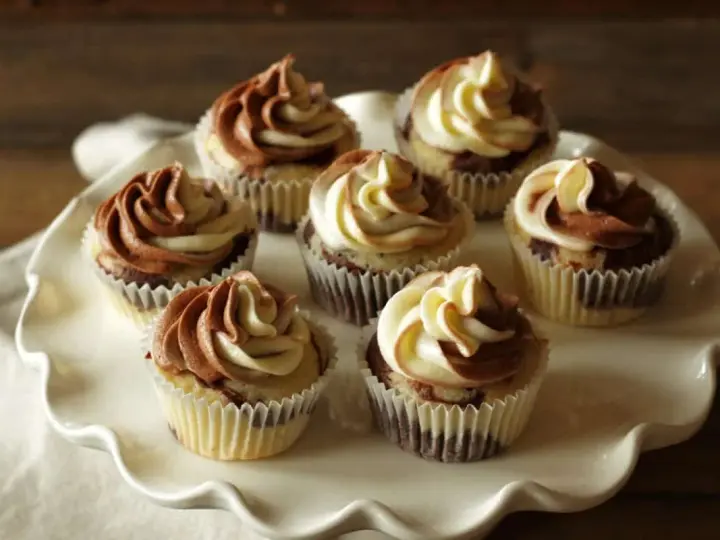 Sieben Marmormuffins mit zweifarbiger Schokoladen-Vanille-Glasur auf einer weißen Tortenplatte. Ein köstliches Marmormuffins Rezept.