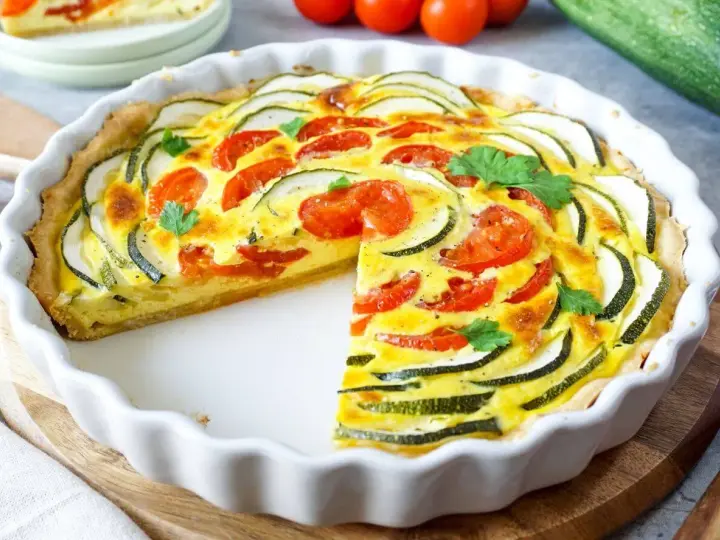 Eine köstliche Zucchini-Quiche mit Tomaten und frischer Petersilie, in einer weißen Quicheform, ein Stück fehlt.