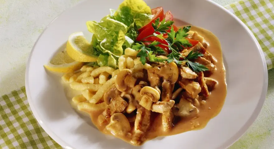 Ein Teller mit geschnetzeltem Fleisch mit Pilzen in cremiger Sauce, dazu Spätzle, Salat und Zitronenspalten.